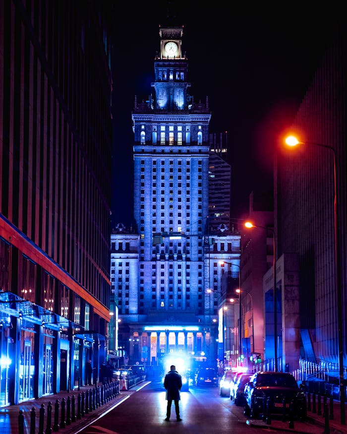 gallery-1 Illuminated Palace of Culture and Science at night with a silhouetted figure and street lights in Warsaw.